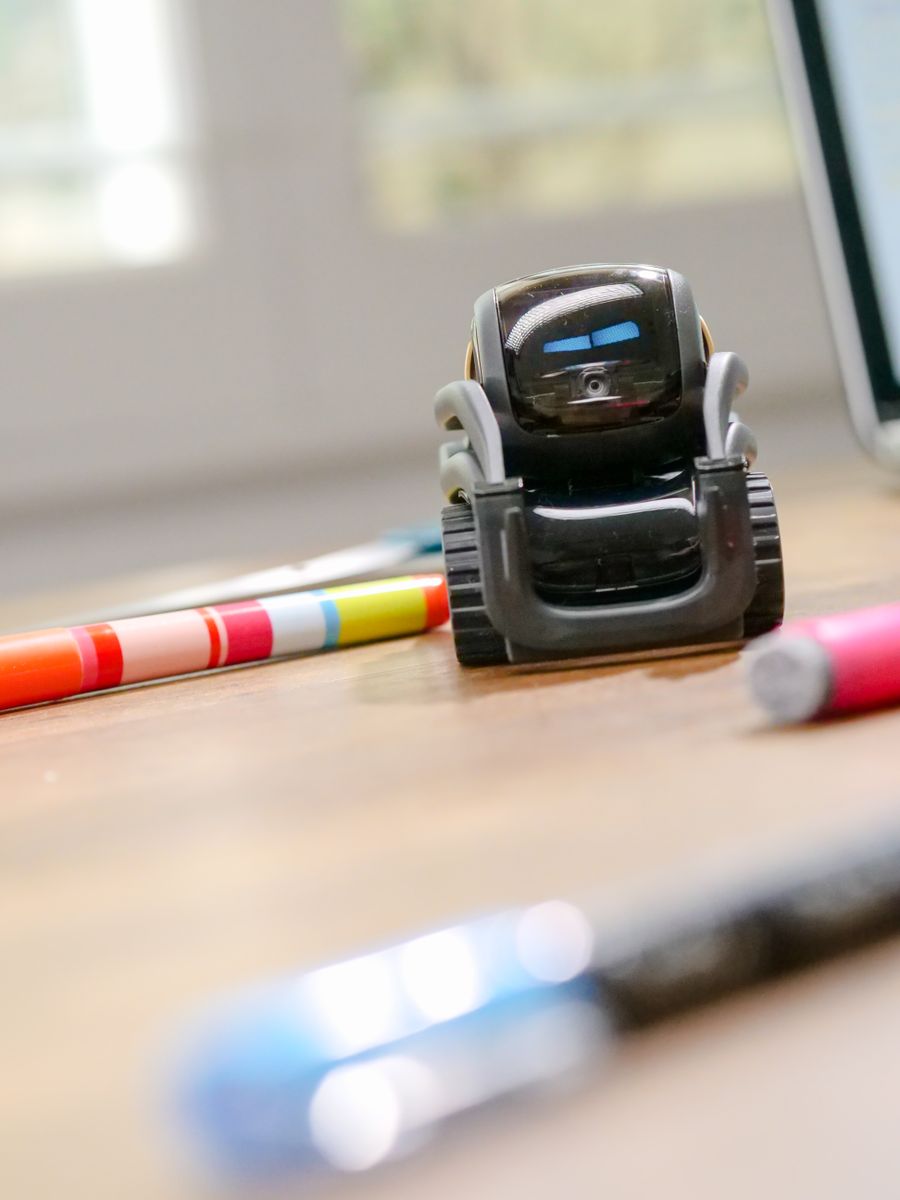 A small robot device sitting on a wooden desk beside pens.